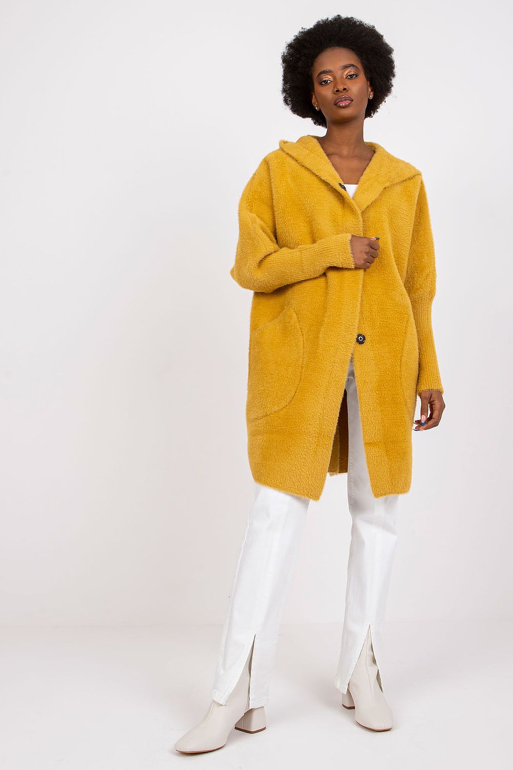 A woman modeling a mustard yellow fuzzy hooded cardigan with front pockets, paired with white pants and white heeled boots, against a plain off-white studio background.
