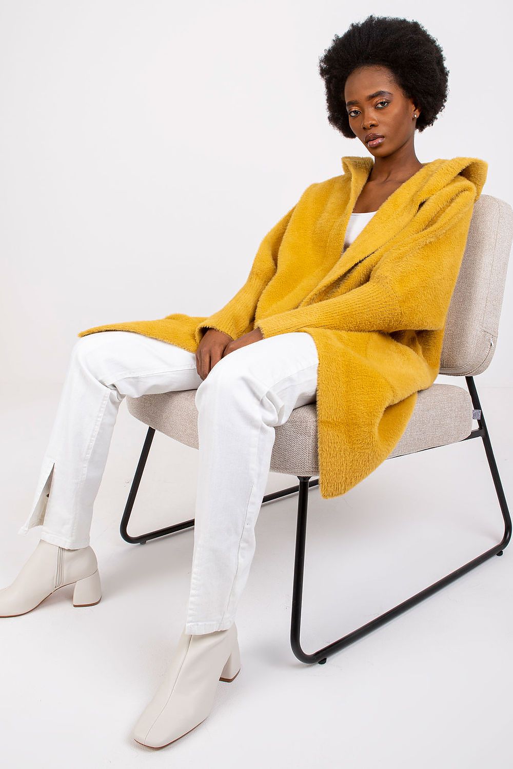 A model sits on a light gray chair wearing a chunky mustard yellow teddy coat, white pants, and off-white heeled boots against a plain white studio background.