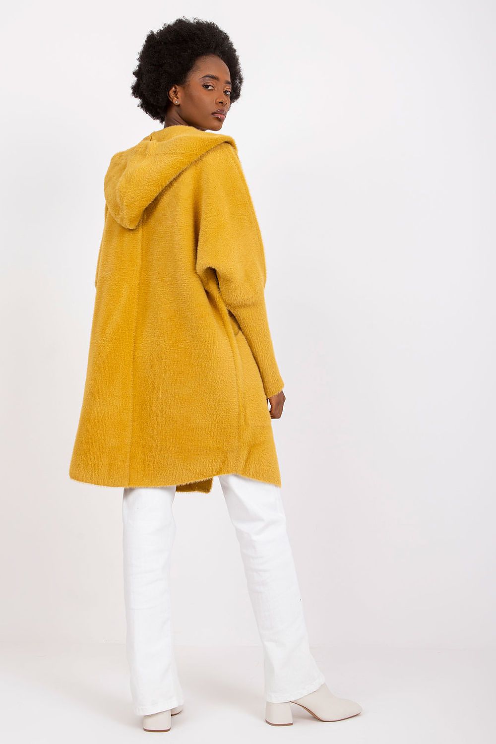 A model wearing a mustard yellow hooded coat, white pants, and white boots, photographed from the back against a plain white studio background.