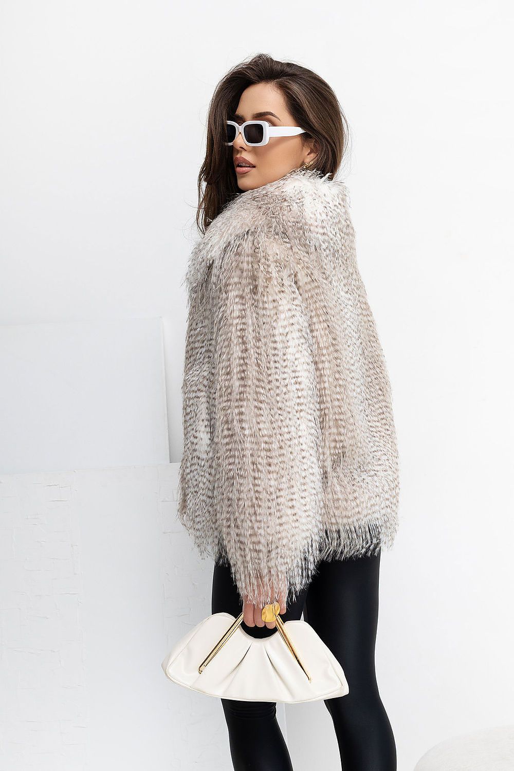 A woman wearing a chunky, textured light beige knit sweater with a high collar, styled with black leggings, white sunglasses, and a white pleated handbag.
