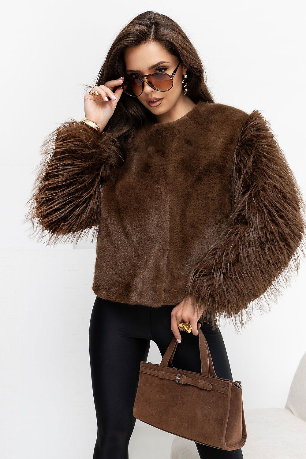 A woman models a brown faux fur cropped jacket with dramatic feathered puff sleeves, paired with black leggings and a matching brown suede handbag against a clean white background.