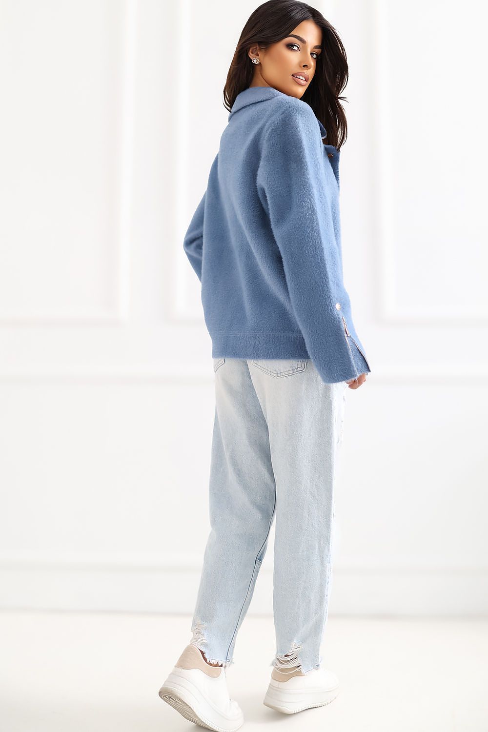 A woman wearing a dusty blue fuzzy knit cardigan, light-wash jeans, and white sneakers, photographed from the back in a bright white studio setting.