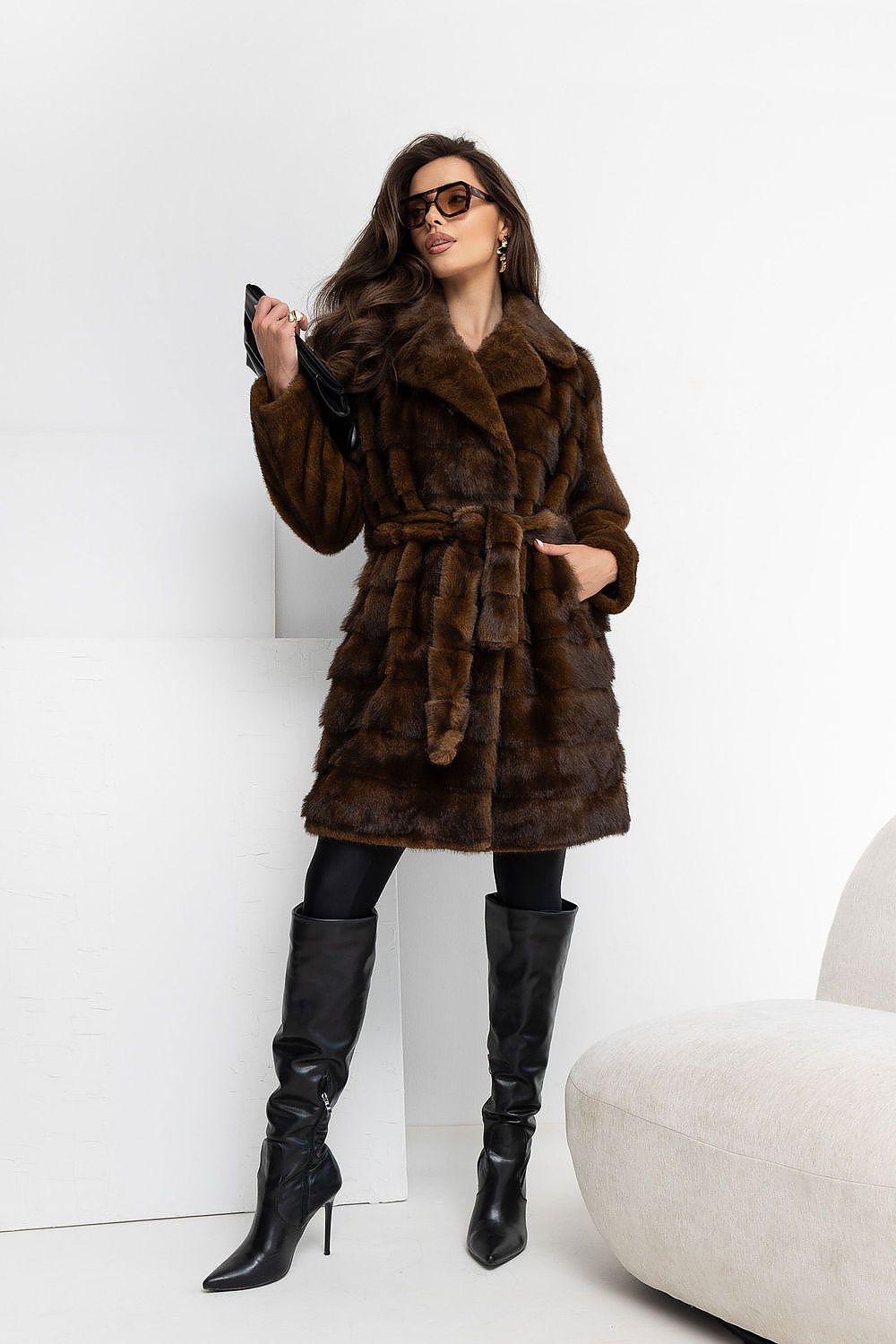 A woman models a dark brown fur coat with a matching belt, styled with black knee-high boots, black pants, and sunglasses in a minimalist white studio setting.