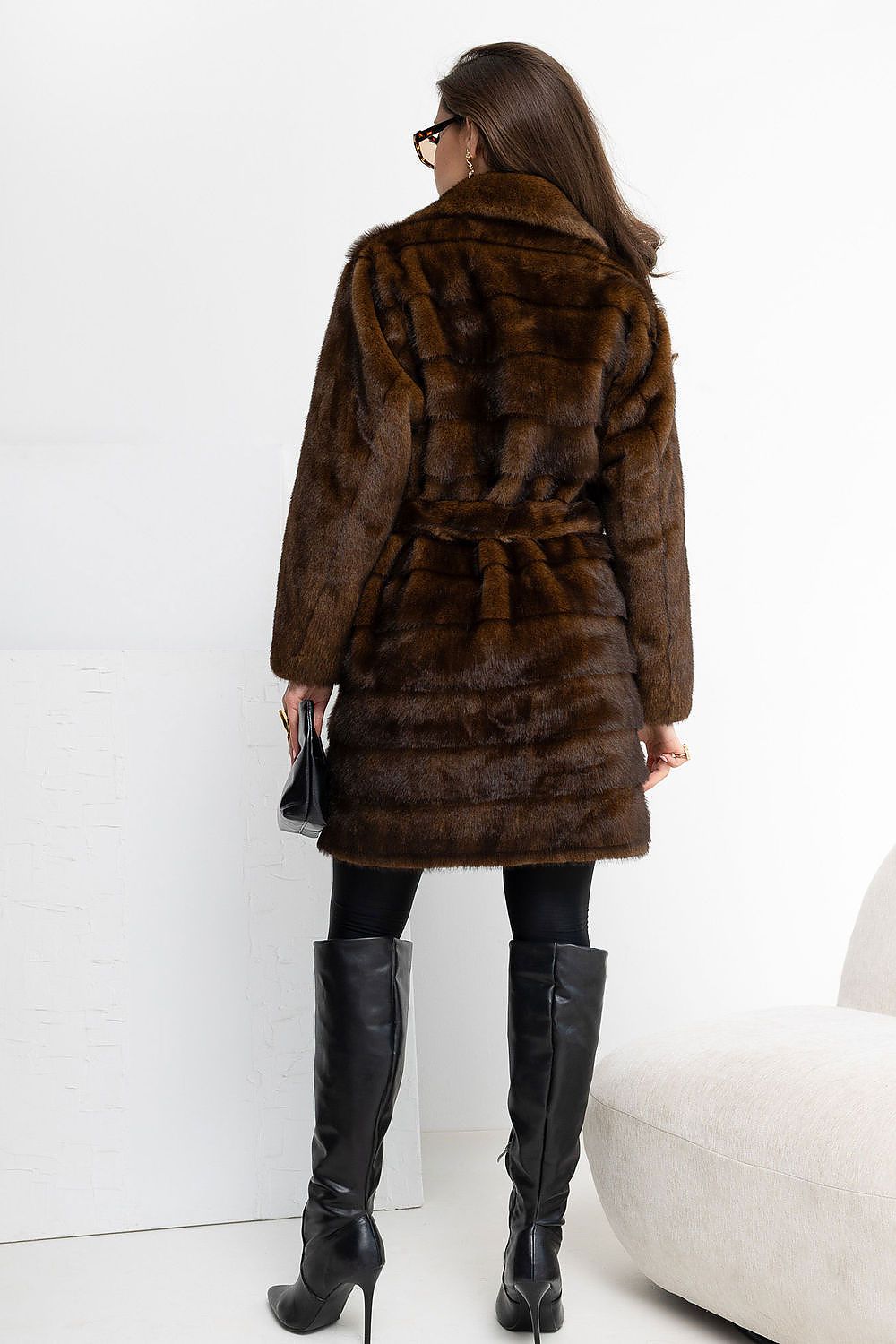 A woman shown from the back wearing a dark brown fur coat with a tie belt, black leggings, and tall black boots, standing in a bright indoor setting.
