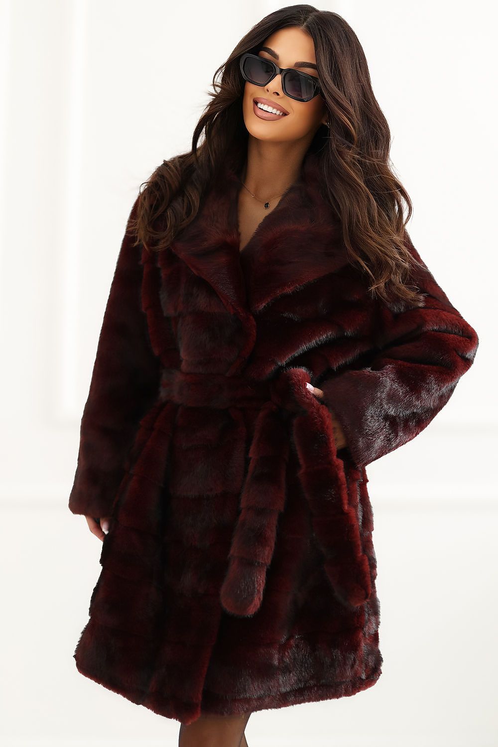 A woman wearing a plush, deep burgundy faux fur coat with a matching belt and sunglasses, posed against a bright white studio background.