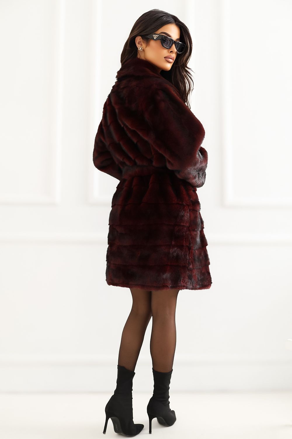 A woman modeling a dark burgundy faux fur coat with a matching belt, paired with black tights and black high-heeled ankle boots, set against a white paneled wall.