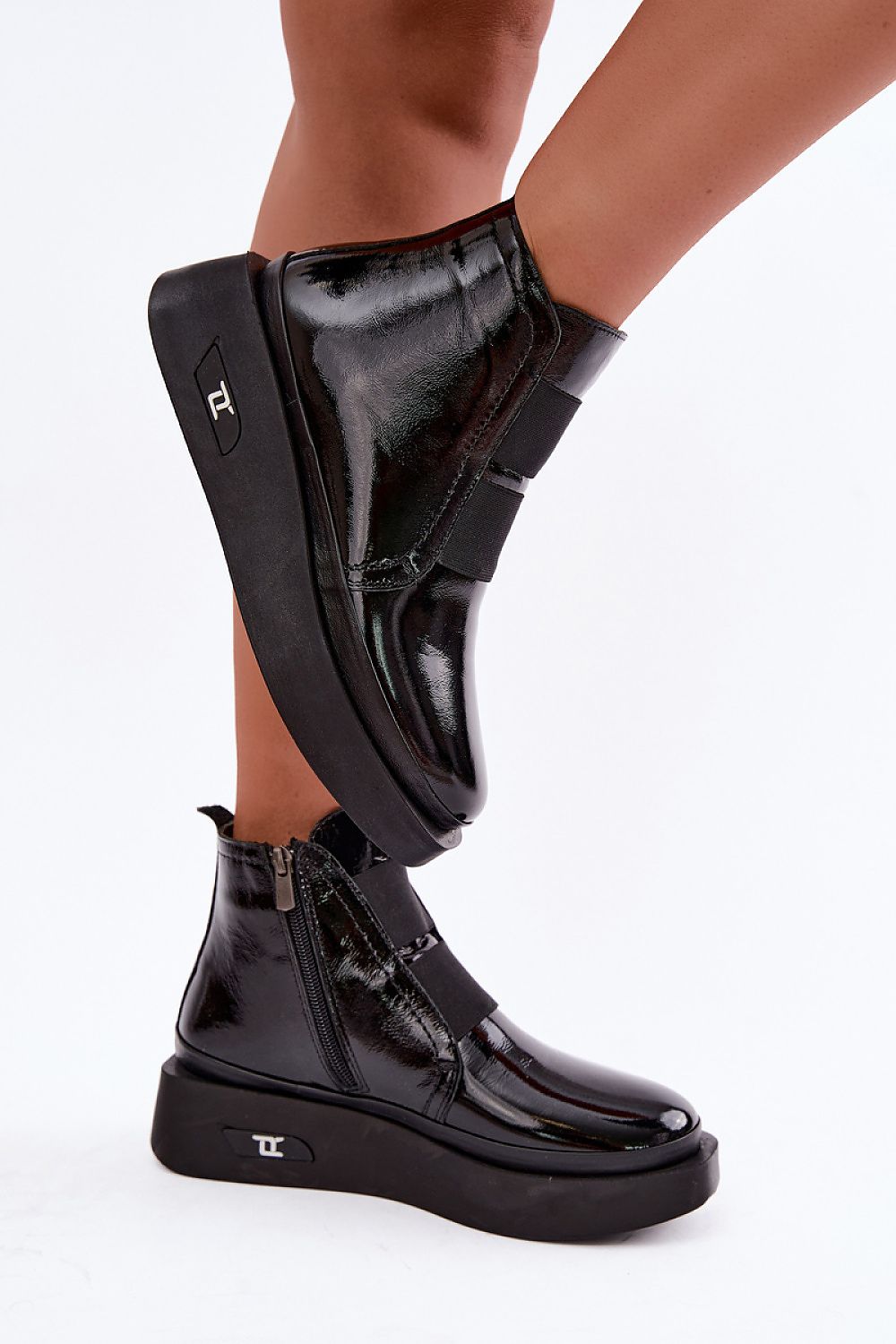A pair of black patent leather ankle boots with chunky platform soles and hook-and-loop strap closures, shown on a model's feet against a white studio background.