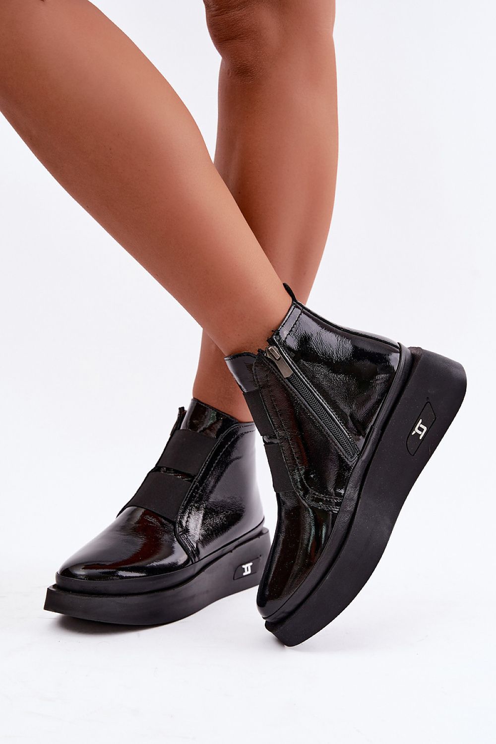 A pair of black patent leather ankle boots with platform soles and side zippers, shown on a model's feet against a white studio background.