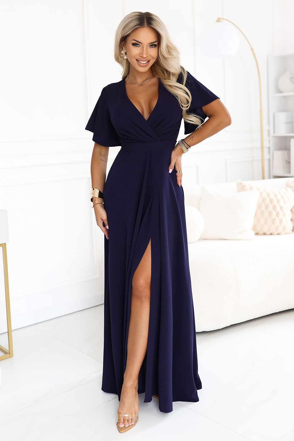 A woman modeling a navy blue maxi dress with short flutter sleeves, a deep V-neckline, and a high thigh slit, standing in a bright, modern living room.