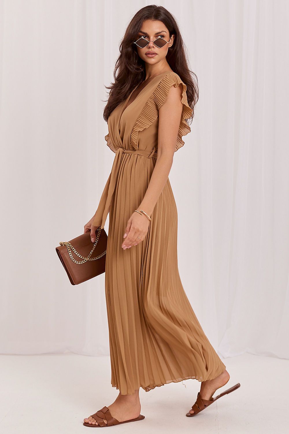 A woman models a tan pleated maxi dress with ruffled shoulder details, holding a brown handbag and wearing brown slide sandals against a white draped backdrop.