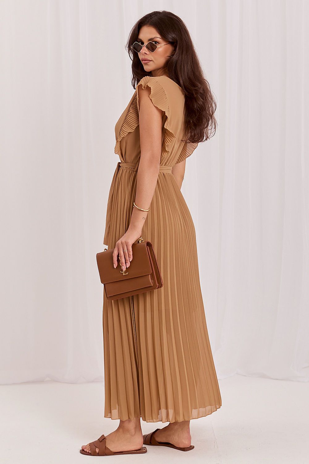 A woman modeling a tan pleated maxi dress with ruffled cap sleeves and a back cutout, holding a matching tan clutch bag against a white draped studio backdrop.