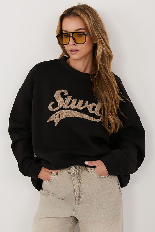 A woman wearing a black crewneck sweatshirt with a beige glittery 'Stuart 81' graphic on the front, paired with light beige jeans and yellow-tinted sunglasses, posing against a light gray wall.