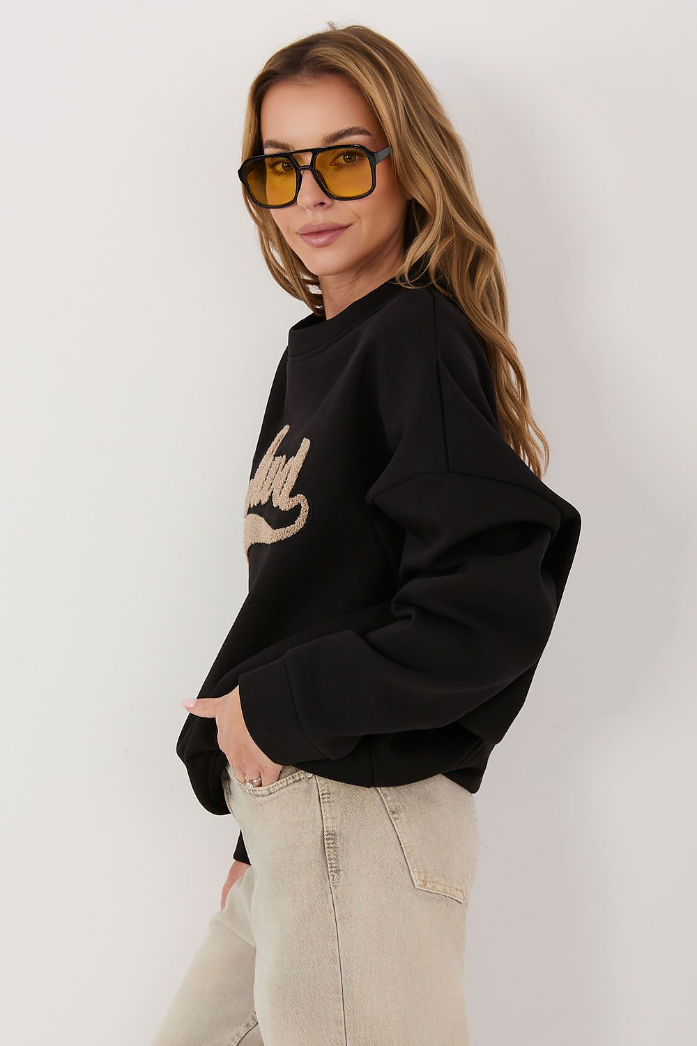 A woman wearing a black cropped sweatshirt with a beige graphic print on the front, paired with light beige jeans and yellow-tinted sunglasses, posing against a plain white studio background.