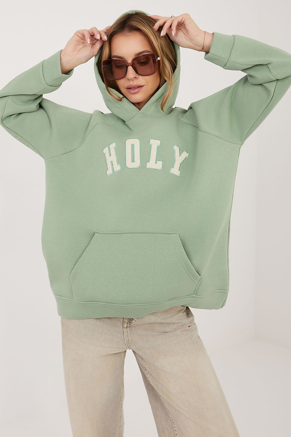 A woman wearing a sage green pullover hoodie with 'HOLY' printed in white arched letters across the chest, paired with beige jeans and sunglasses, against a plain light background.