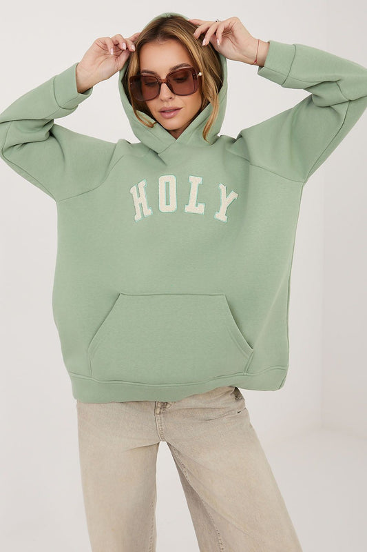 A woman wearing a sage green pullover hoodie with 'HOLY' printed in white arched letters across the chest, paired with beige jeans and sunglasses, against a plain light background.