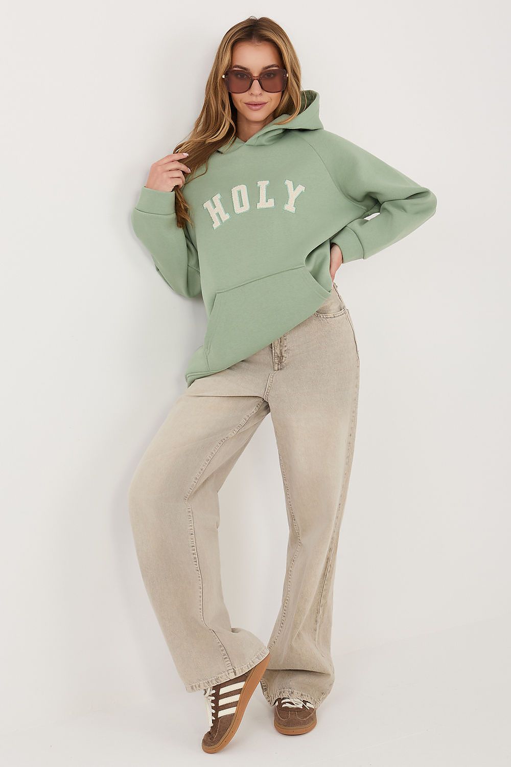 A woman modeling a sage green pullover hoodie with 'HOLY' printed in white arched letters across the chest, paired with beige wide-leg jeans and brown sneakers, against a plain light gray studio background.