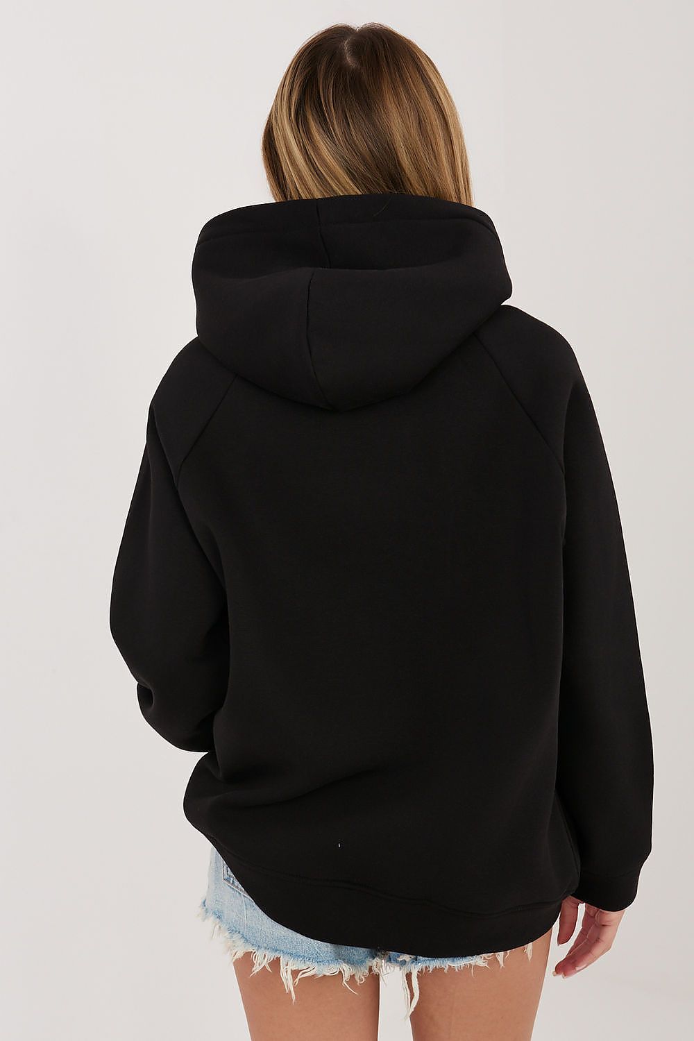 A woman viewed from the back wearing a black pullover hoodie and light blue denim shorts against a plain light gray studio background.