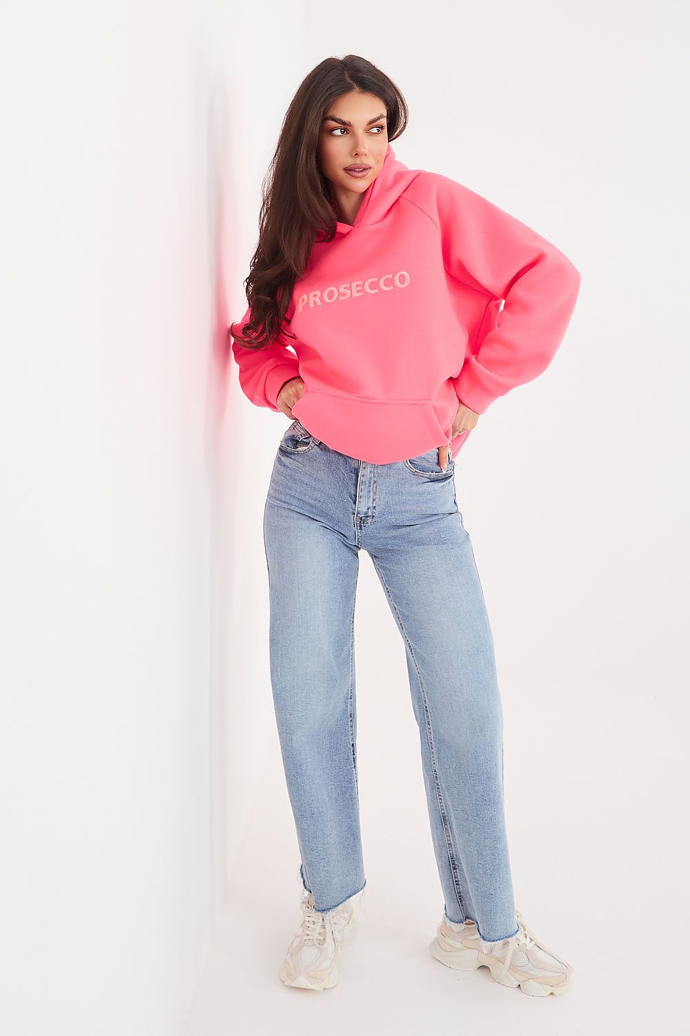 A woman posing in a bright pink cropped hoodie with 'PROSECCO' printed in white across the chest, paired with light-wash blue jeans and white sneakers, leaning against a white wall.