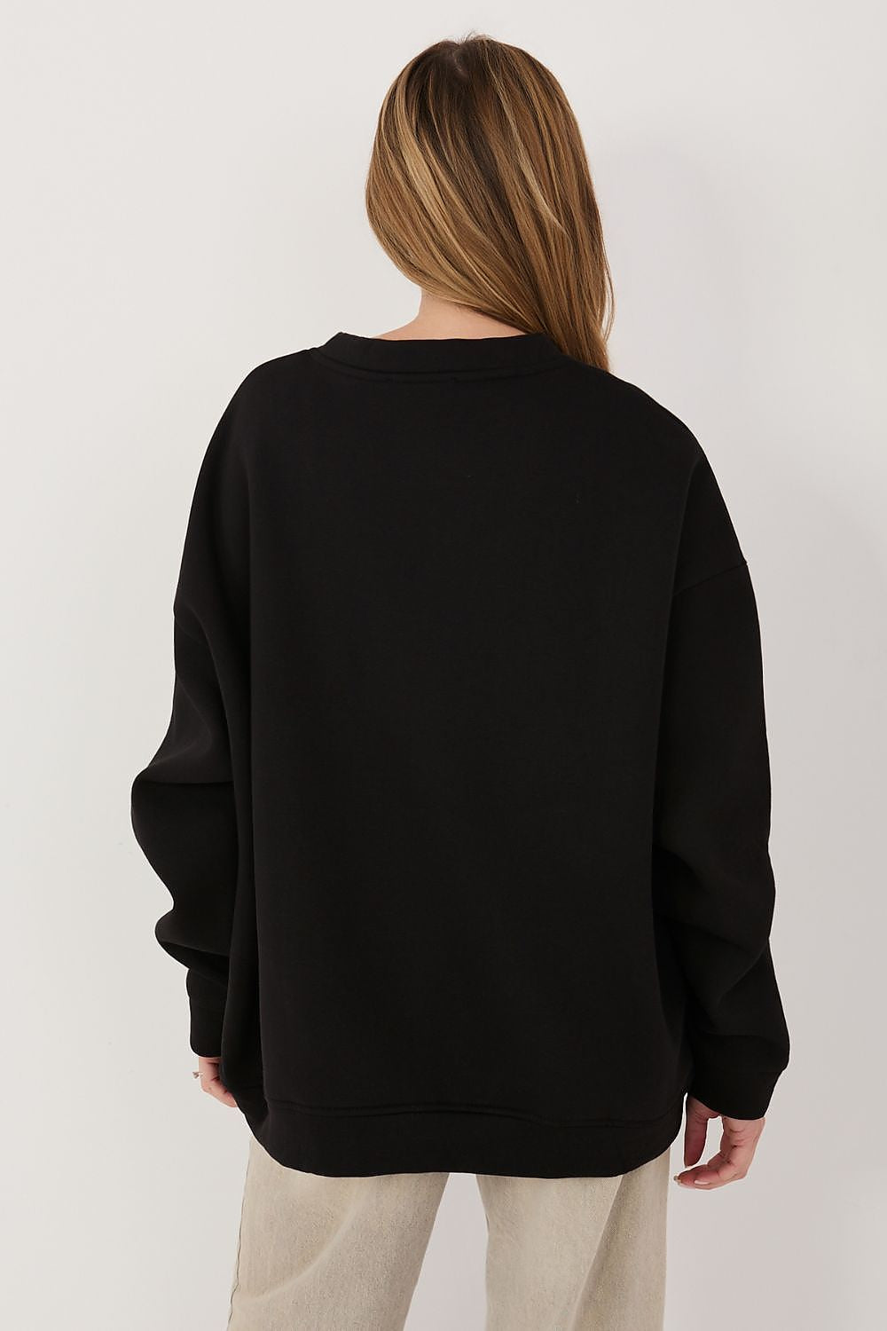 A woman viewed from the back wearing an oversized black crewneck sweatshirt and light beige pants, set against a plain light gray studio background.