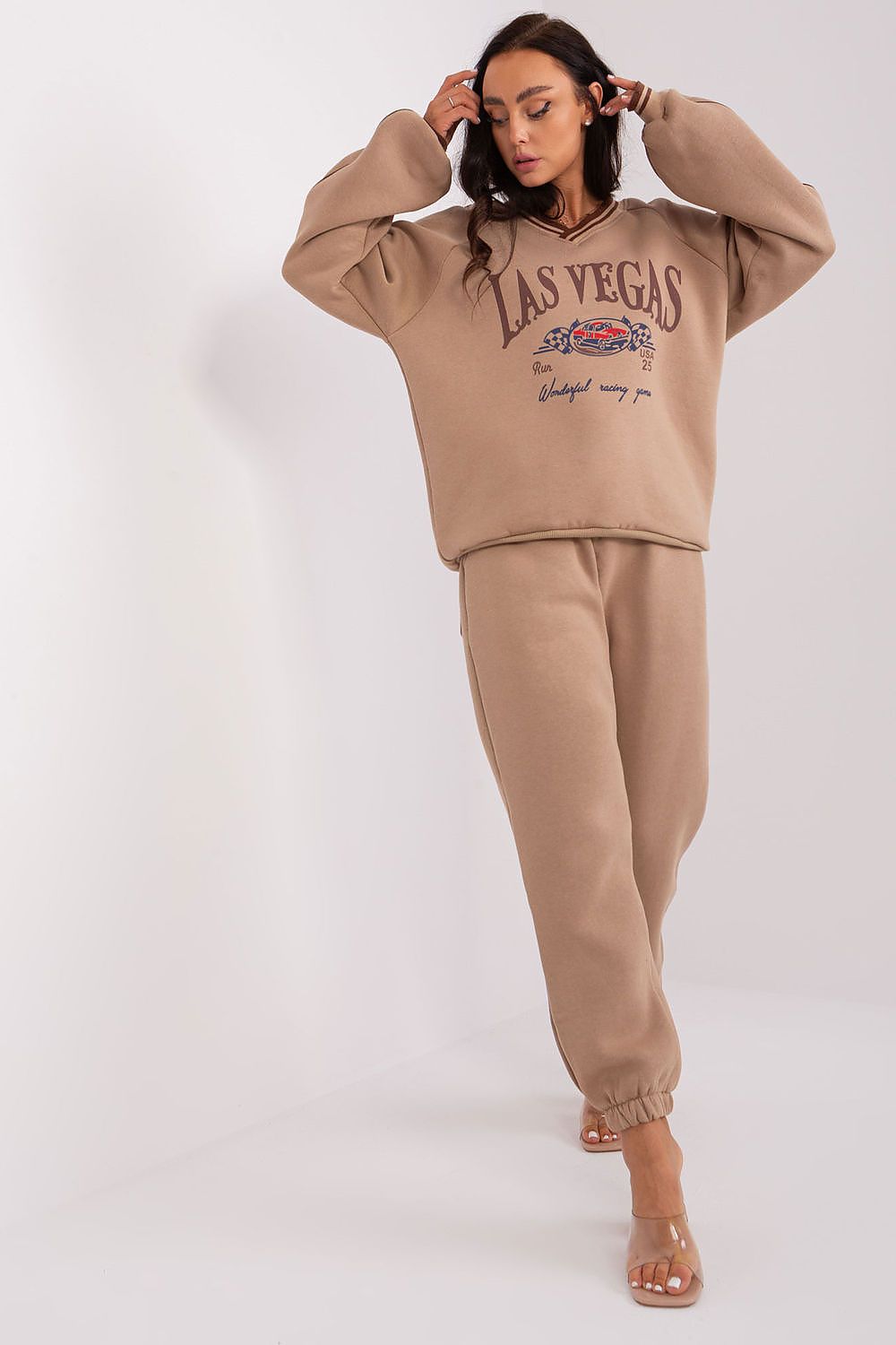 A woman modeling a matching beige lounge set consisting of a cropped sweatshirt with 'LAS VEGAS' text and jogger pants, photographed against a plain light background.