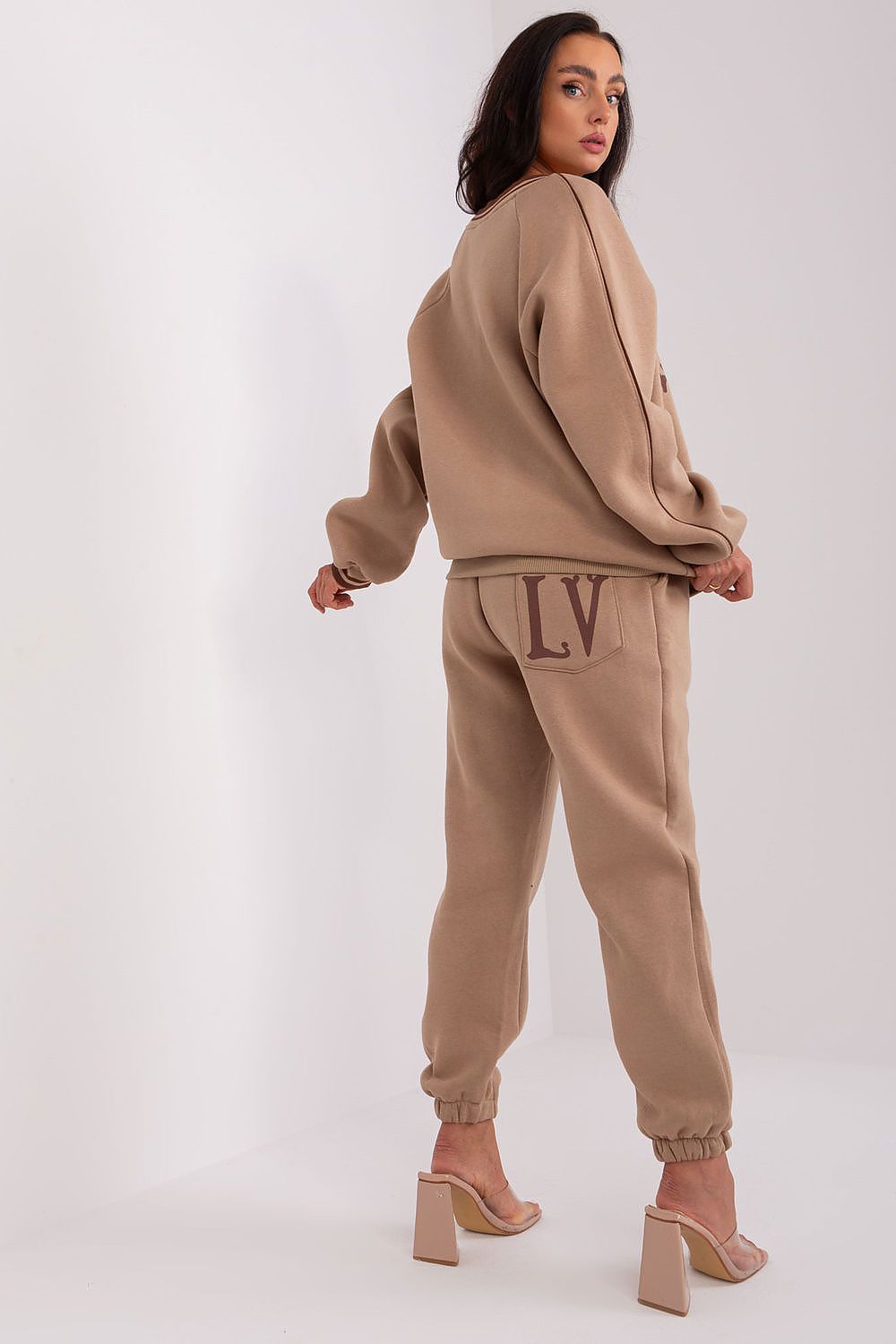 A woman modeling a matching beige lounge set consisting of a cropped sweatshirt and jogger pants with 'LV' embroidered on the back pocket, shown from a rear three-quarter angle against a white studio background.