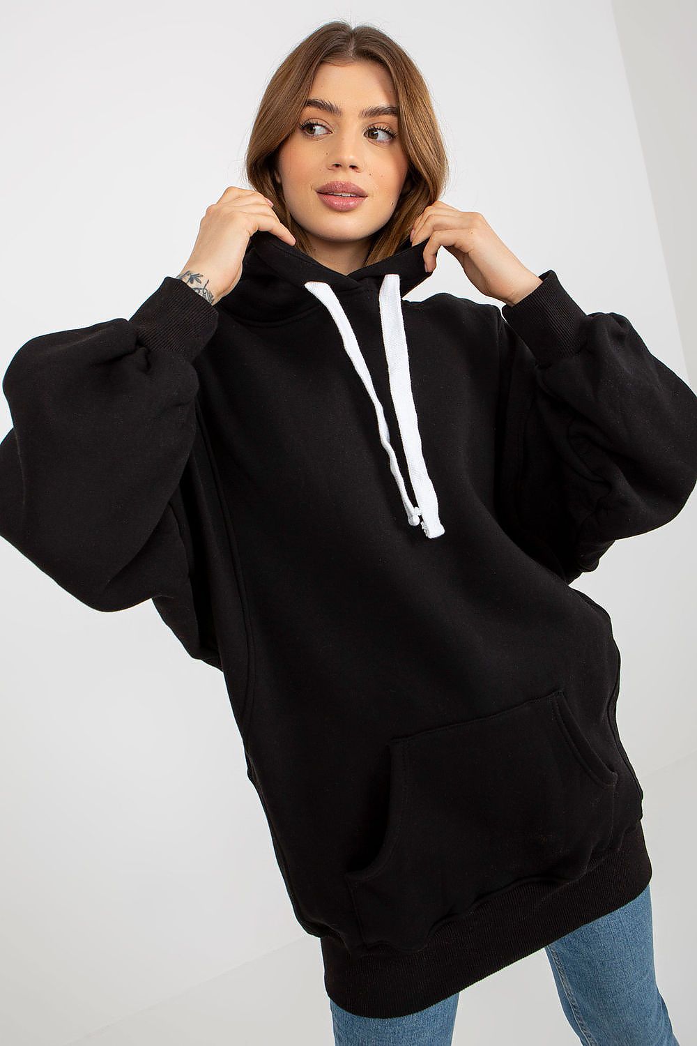 A woman wearing an oversized black pullover hoodie with white drawstrings, paired with blue jeans, posing against a light gray studio background.
