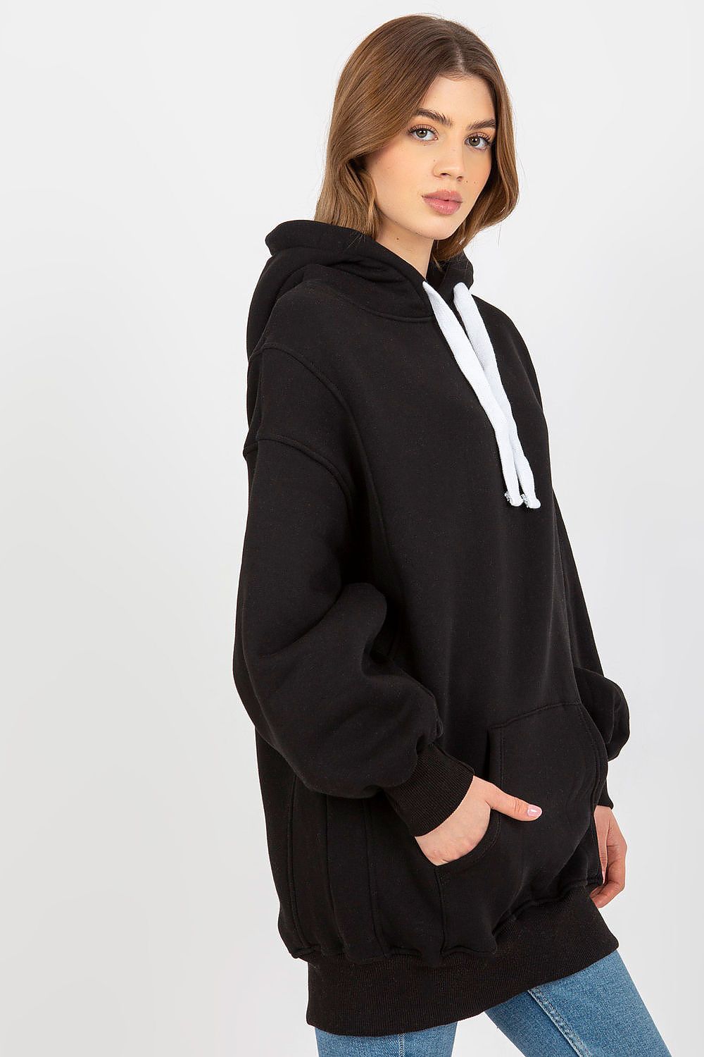 A woman wearing an oversized black pullover hoodie with white drawstrings, paired with blue jeans, photographed against a light gray studio background.