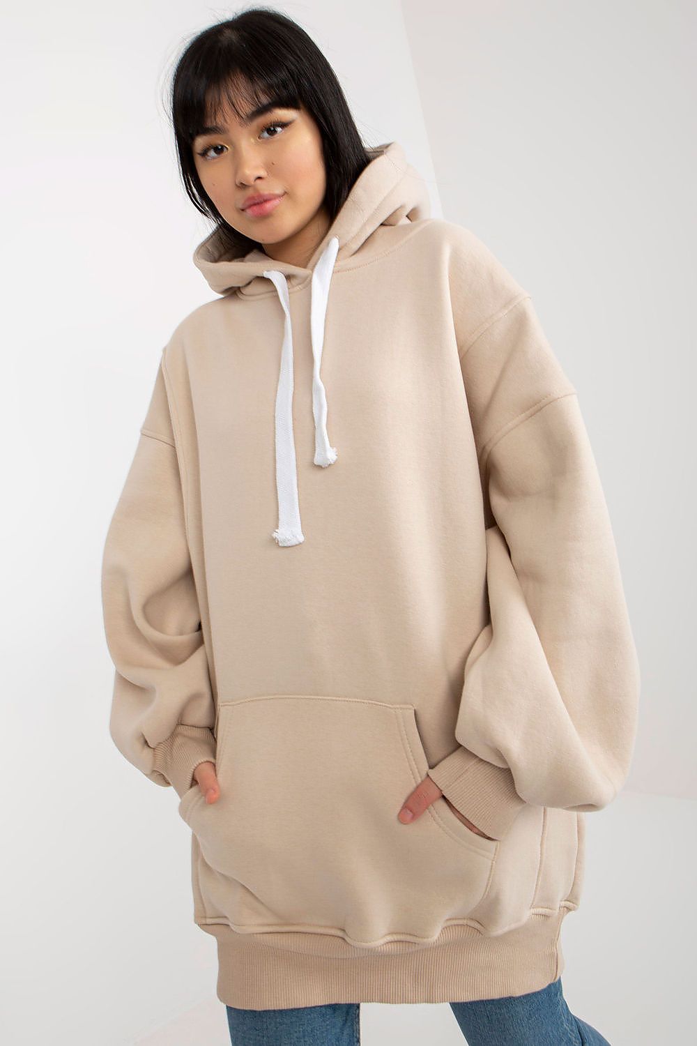 A woman wearing an oversized beige hoodie with white drawstrings and a front kangaroo pocket, paired with blue jeans, against a plain light gray studio background.
