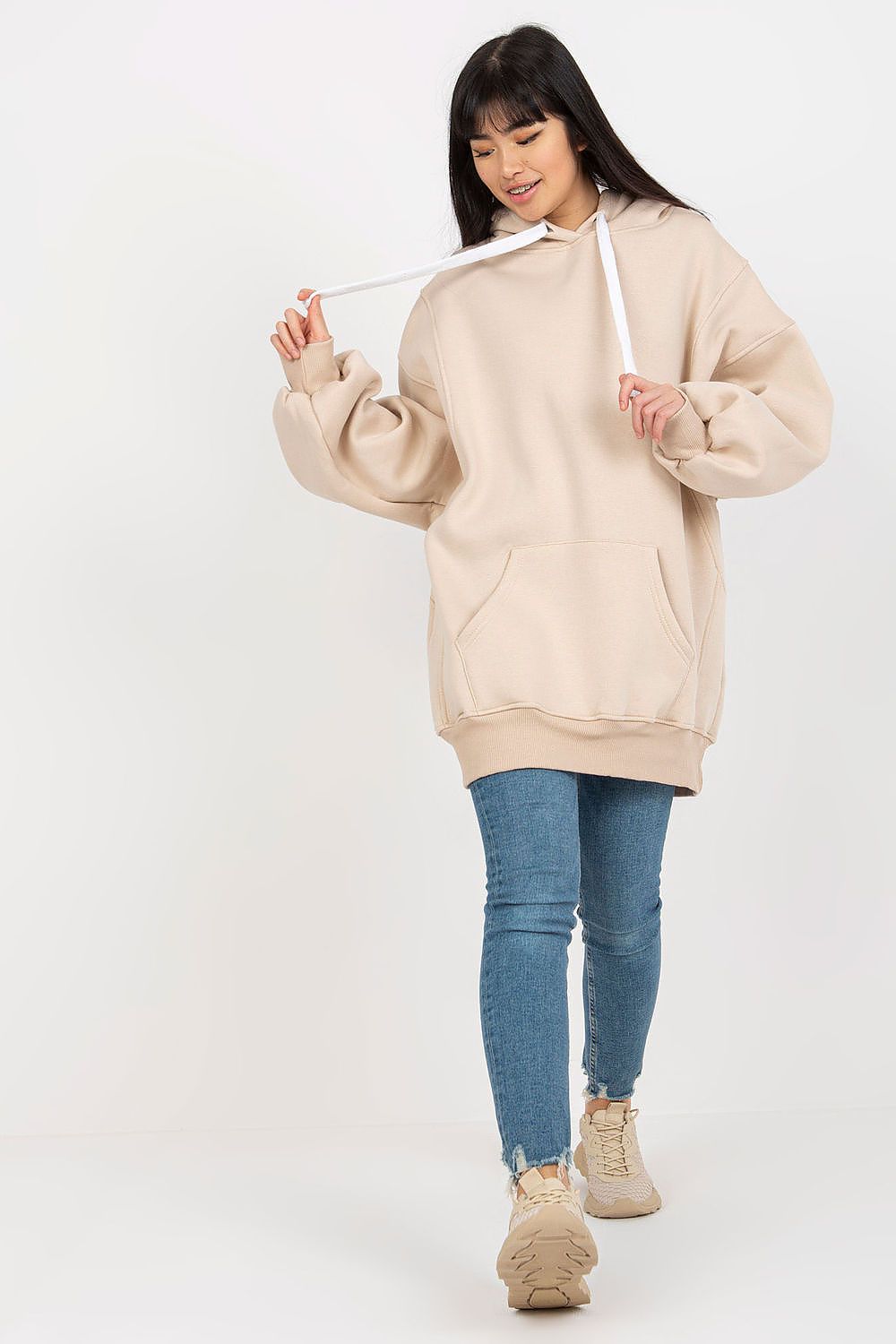 A woman modeling an oversized beige hoodie with white drawstrings, paired with blue skinny jeans and beige sneakers, against a plain white studio background.