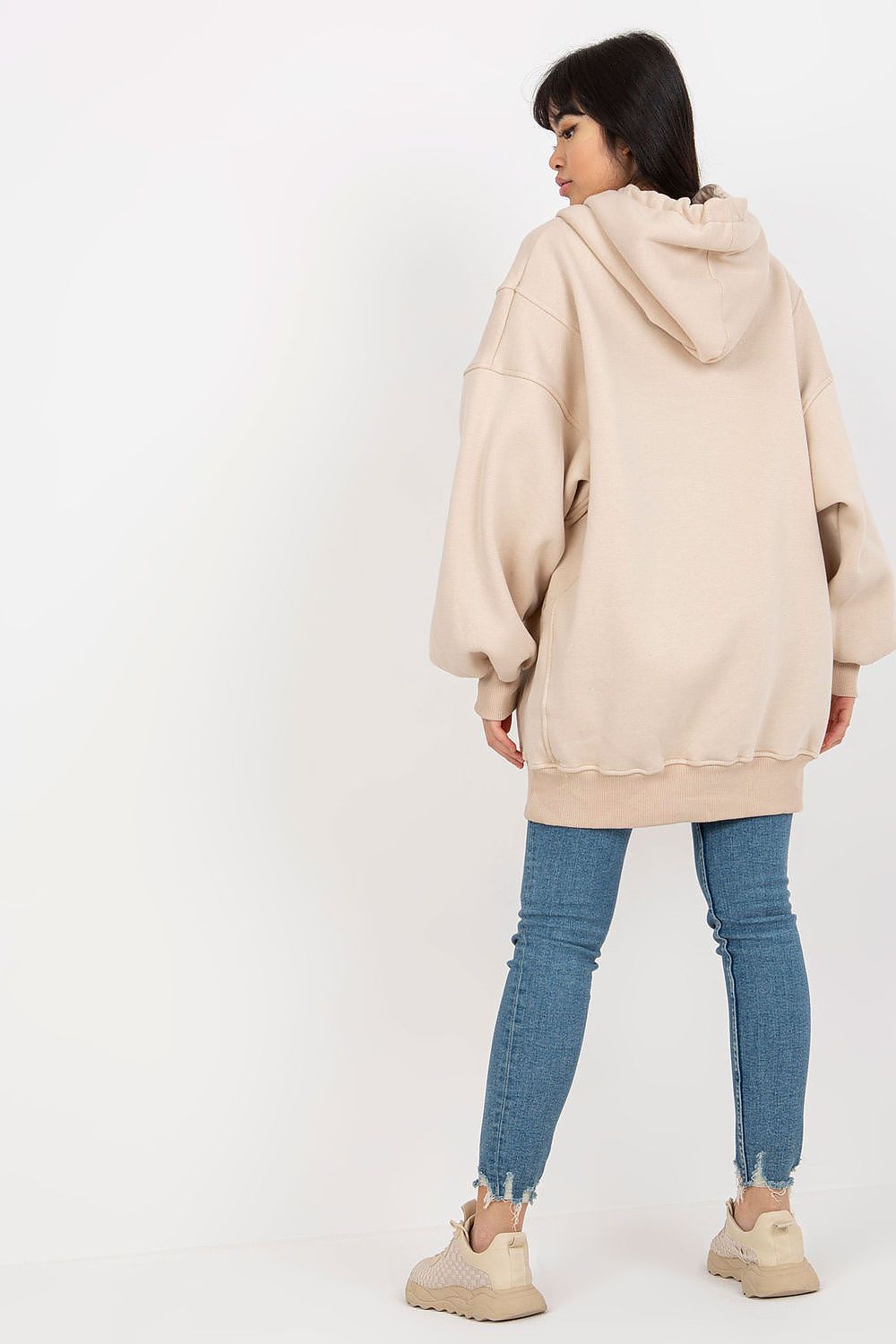 A woman shown from the back wearing an oversized light beige hoodie, blue skinny jeans, and beige sneakers against a plain white studio background.