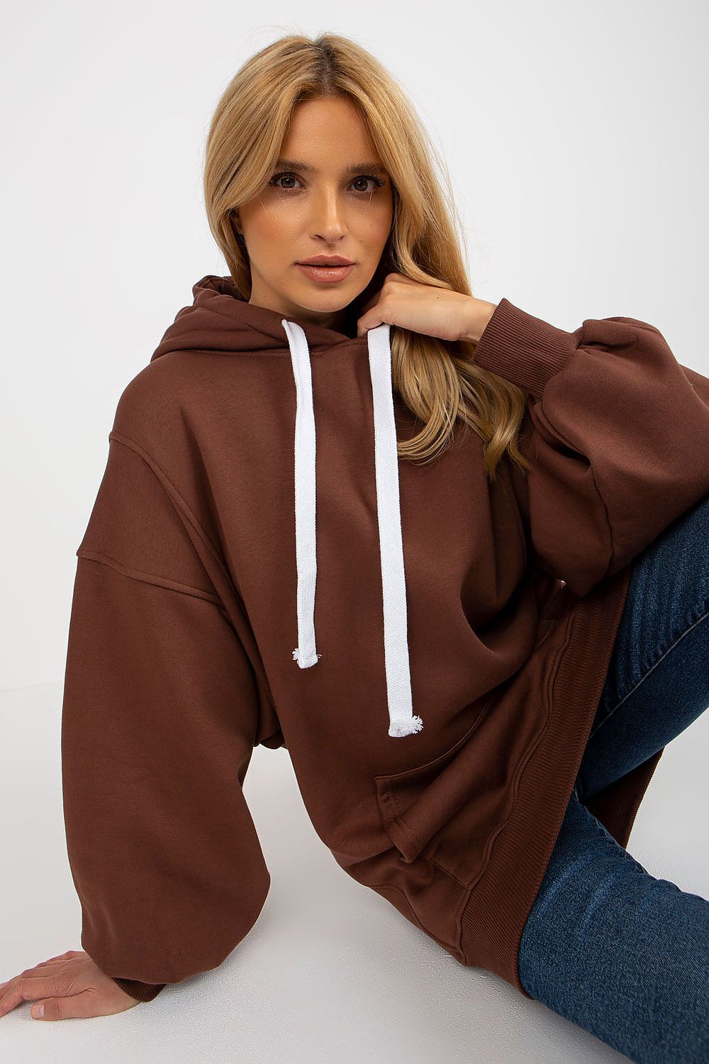A woman wearing an oversized brown hoodie with white drawstrings and blue jeans, posing against a clean white studio background.