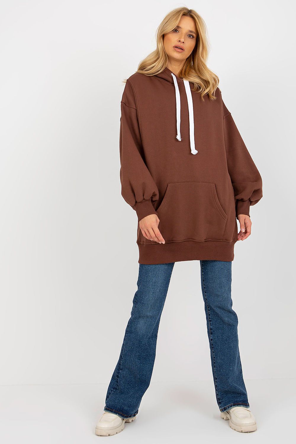 A woman modeling an oversized brown pullover hoodie with white drawstrings, paired with blue jeans and white sneakers, against a plain white studio background.