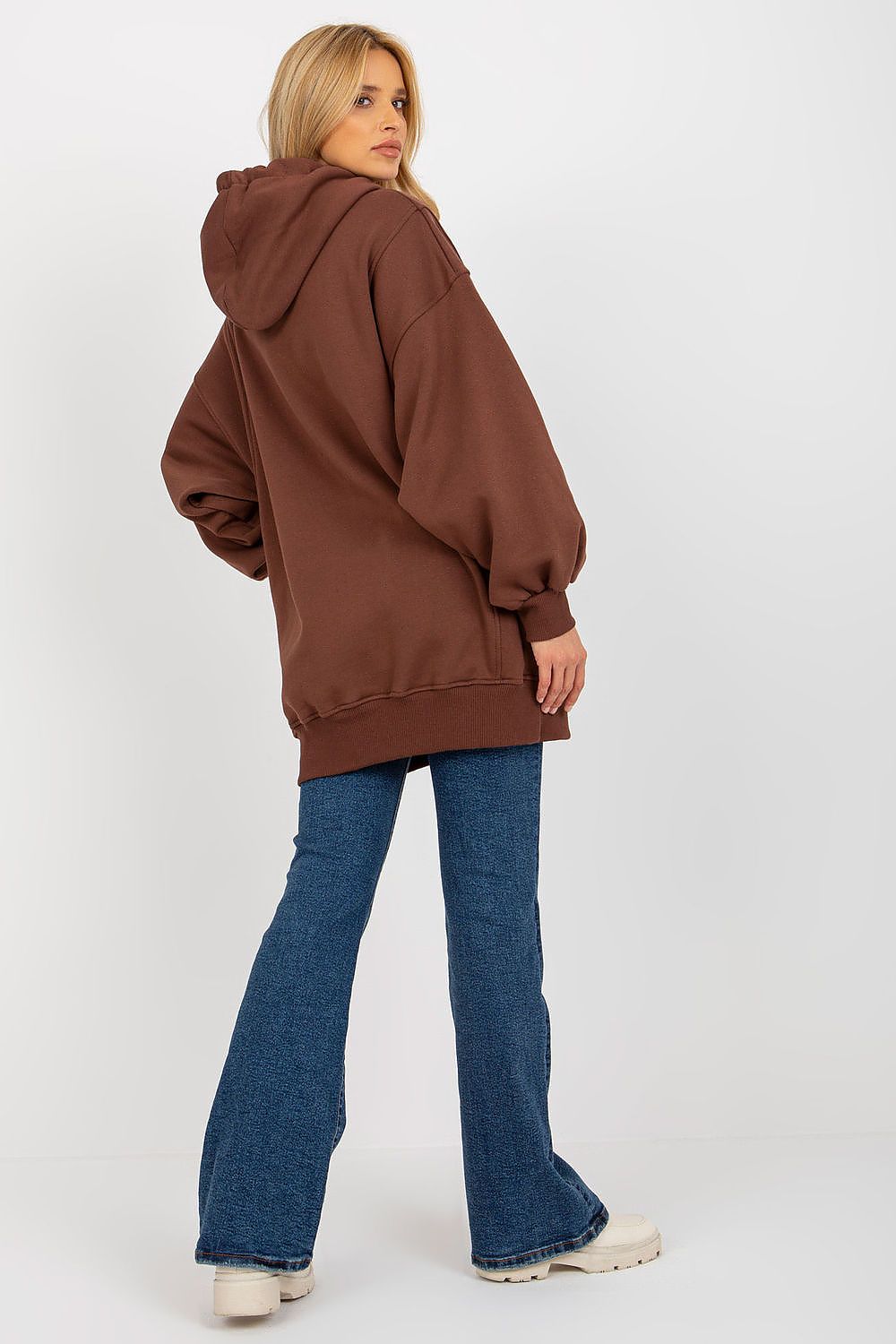 A woman wearing an oversized brown hoodie, blue flare jeans, and white sneakers, photographed from the back against a plain light gray studio background.