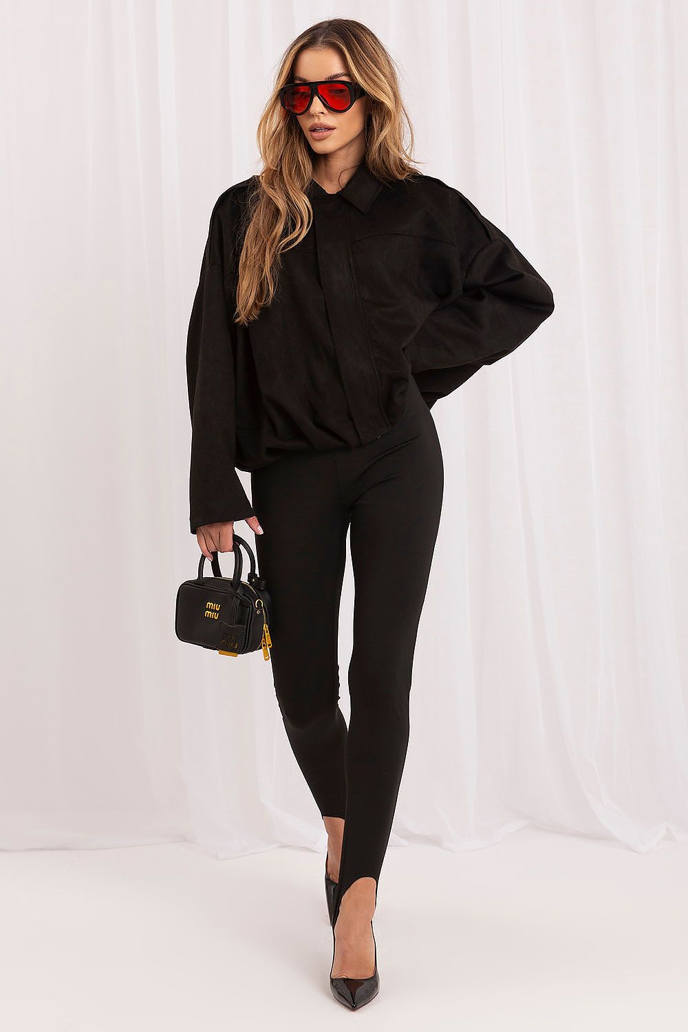 A woman modeling a black long-sleeve collared shirt, black skinny pants, black pointed-toe heels, and red-tinted sunglasses, holding a small black handbag with gold hardware against a white draped studio backdrop.