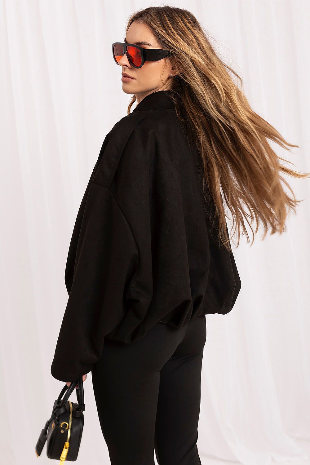 A woman with long blonde hair wearing a black oversized jacket, black pants, and black sunglasses with red-orange lenses, holding a small black handbag against a white draped background.