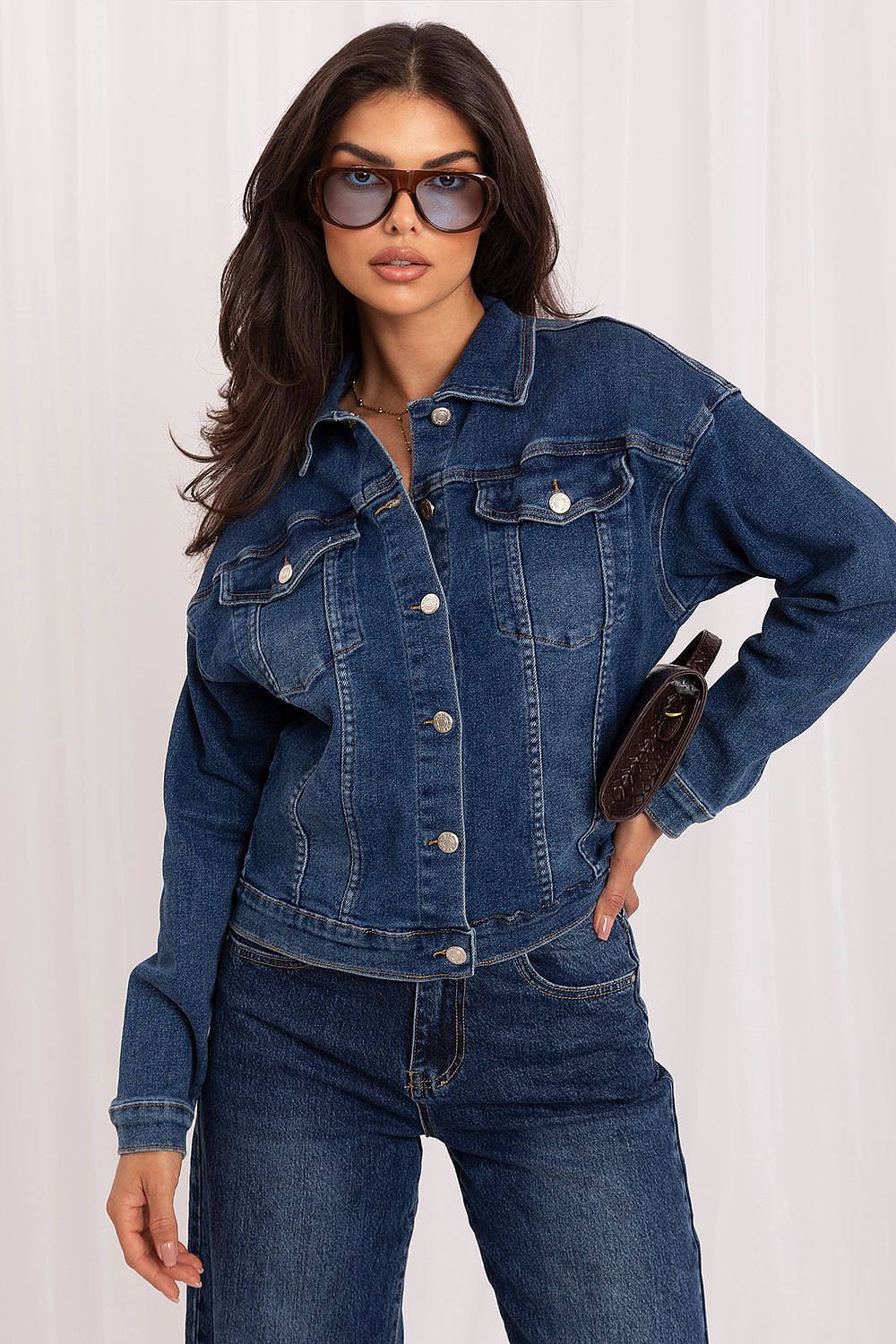 A woman modeling a dark blue denim jacket with matching dark blue jeans, styled with sunglasses and a brown clutch, against a white curtain background.
