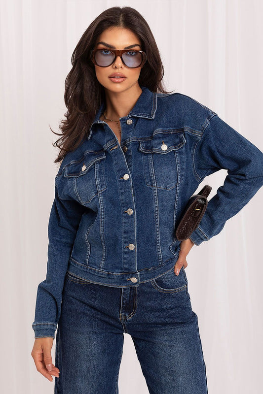 A woman modeling a dark blue denim jacket with matching dark blue jeans, styled with sunglasses and a brown clutch, against a white curtain background.