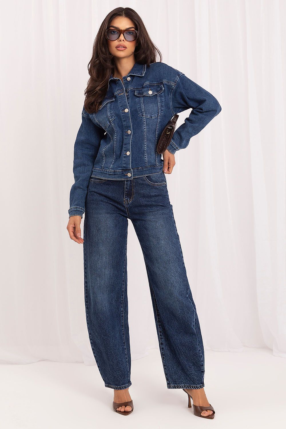 A woman modeling a dark blue denim jacket and matching wide-leg denim jeans, styled with brown heeled sandals and sunglasses against a white draped studio backdrop.