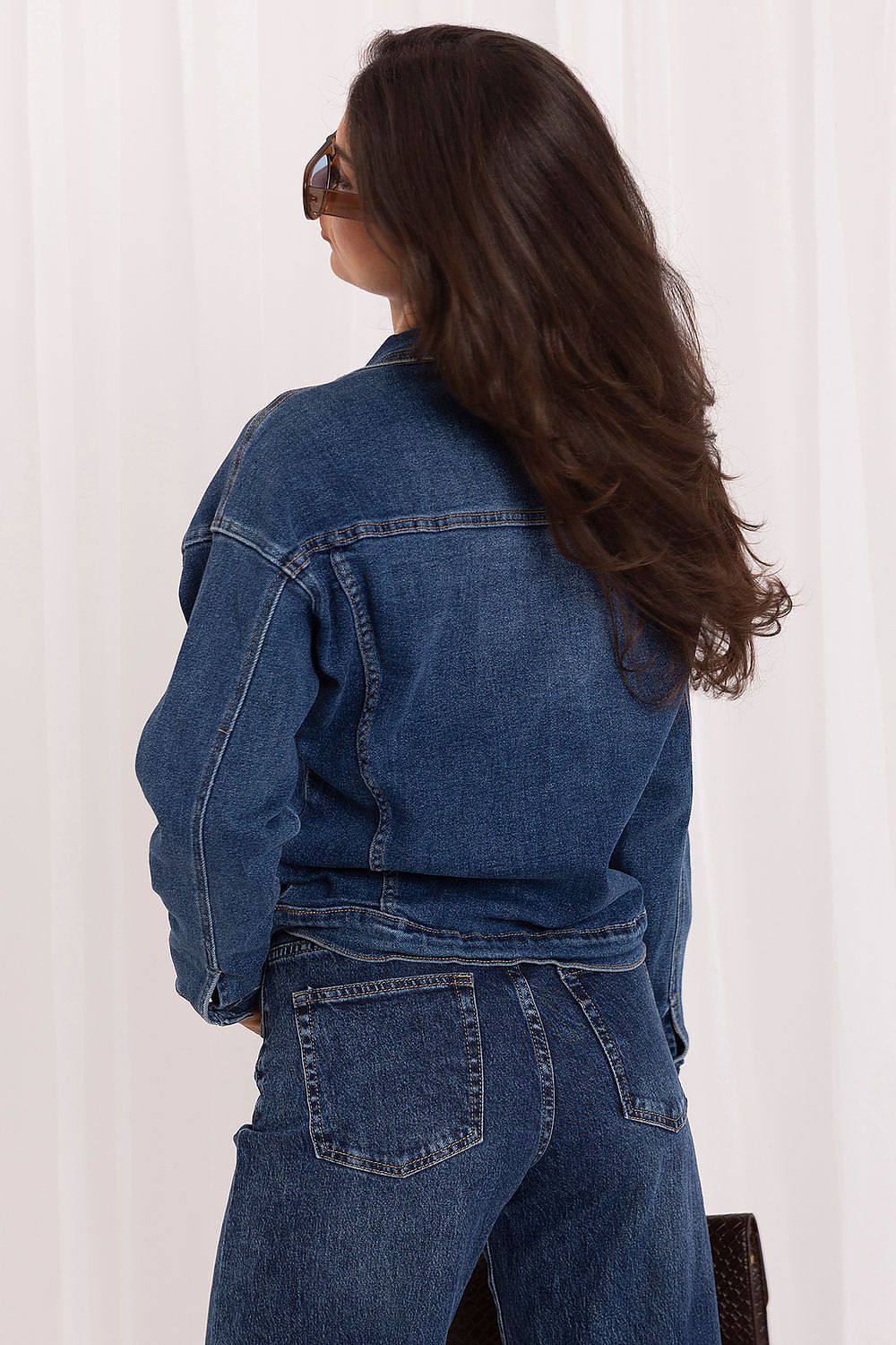 A woman with long brown hair is shown from the back wearing a blue denim jacket and matching blue denim jeans against a white curtain background.