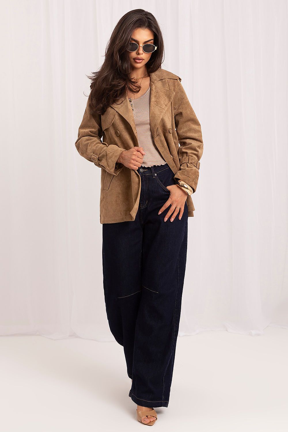 A woman modeling a tan suede jacket over a beige top and dark wide-leg jeans, standing against a white draped studio backdrop.