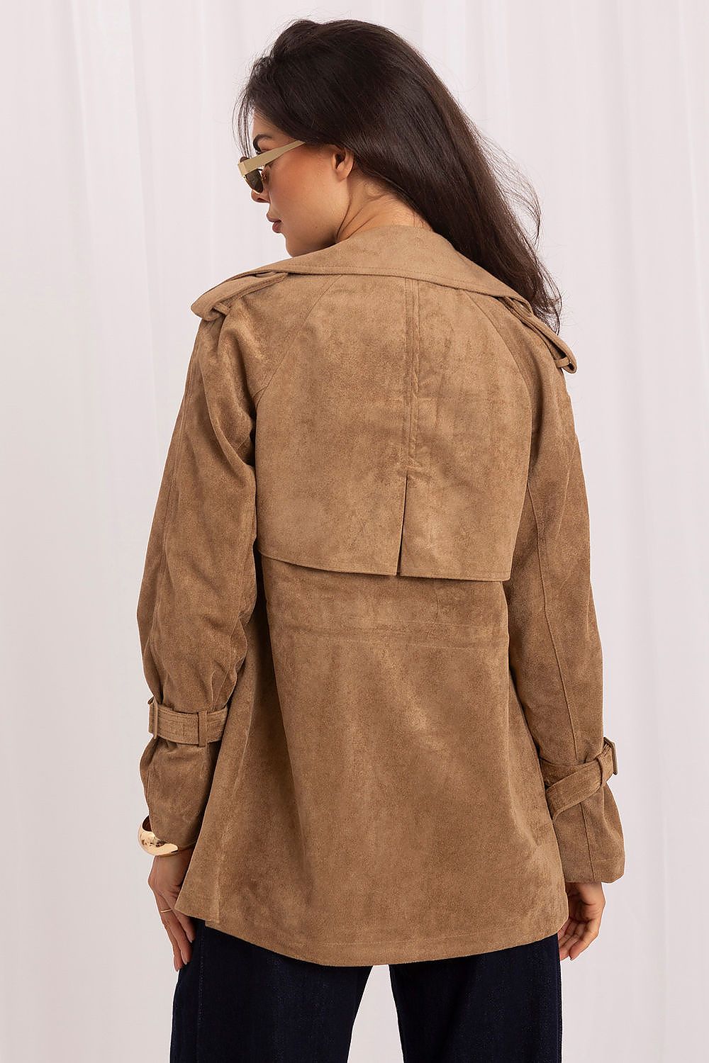 A woman shown from the back wearing a tan suede trench coat with dark pants, photographed against a white curtain backdrop.