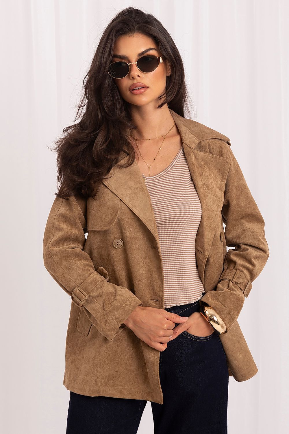 A woman modeling a tan suede-like double-breasted blazer over a beige and white striped top with dark blue jeans, set against a white curtain background.