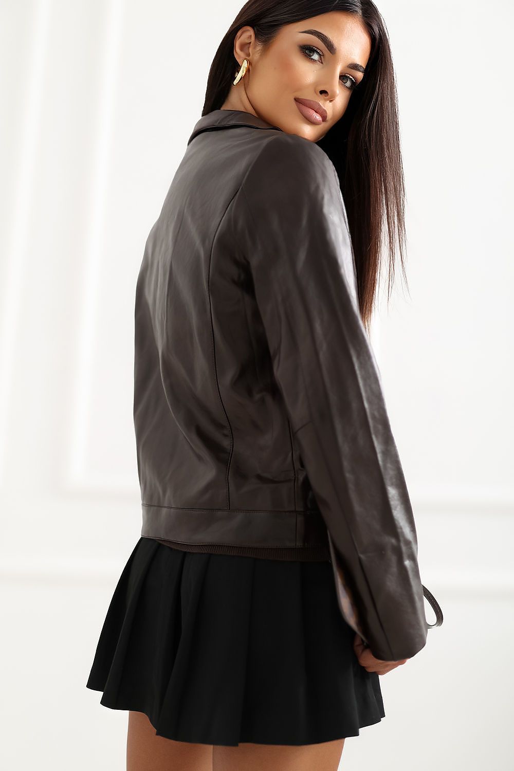 A woman wearing a dark brown faux-leather jacket and a black pleated mini skirt, photographed from the back against a white paneled wall.
