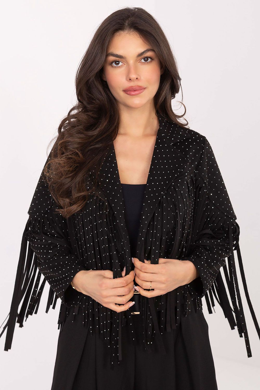 A woman modeling a black fringed cardigan jacket embellished with small silver studs, worn over a black top and black skirt against a plain white studio background.