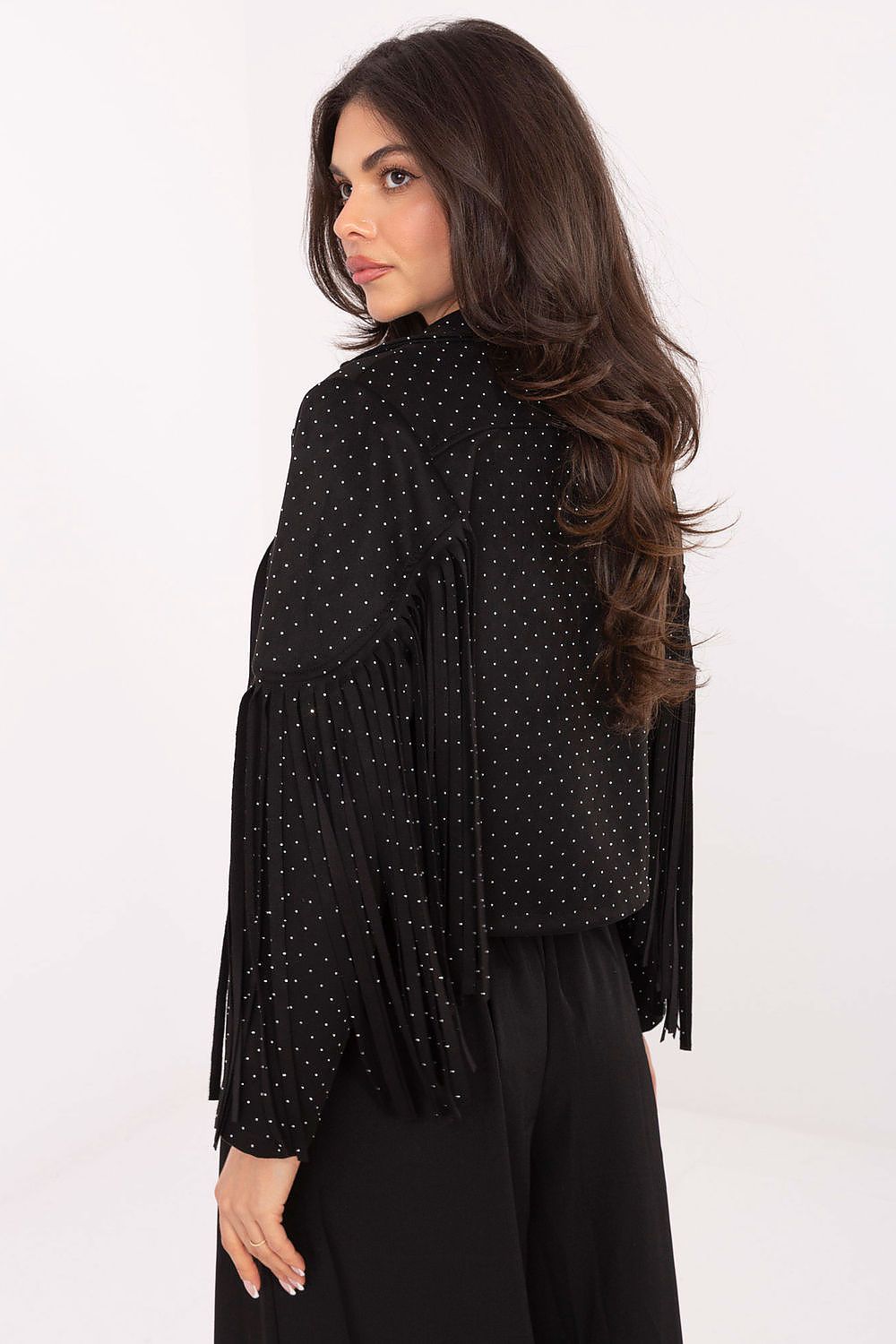 A woman wearing a black long-sleeve fringed top with small white polka dots, shown from a three-quarter back view against a plain white studio background.