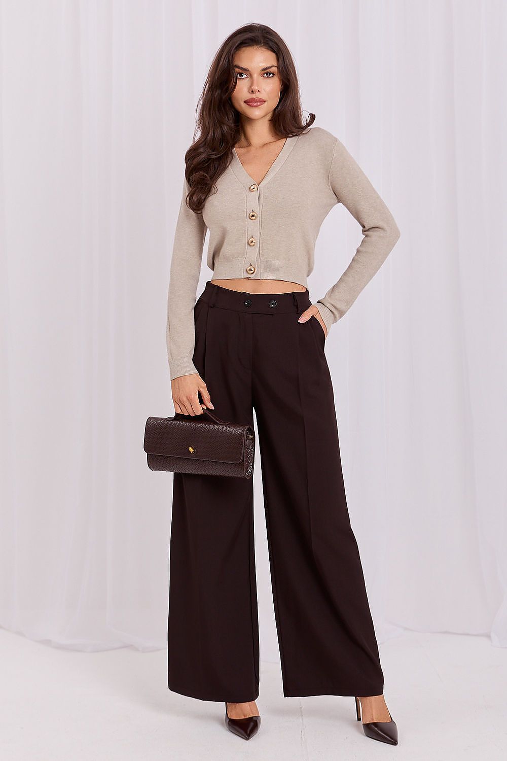 A woman models a beige button-up cardigan and dark brown wide-leg trousers, holding a dark brown crocodile-embossed clutch bag against a white draped studio backdrop.
