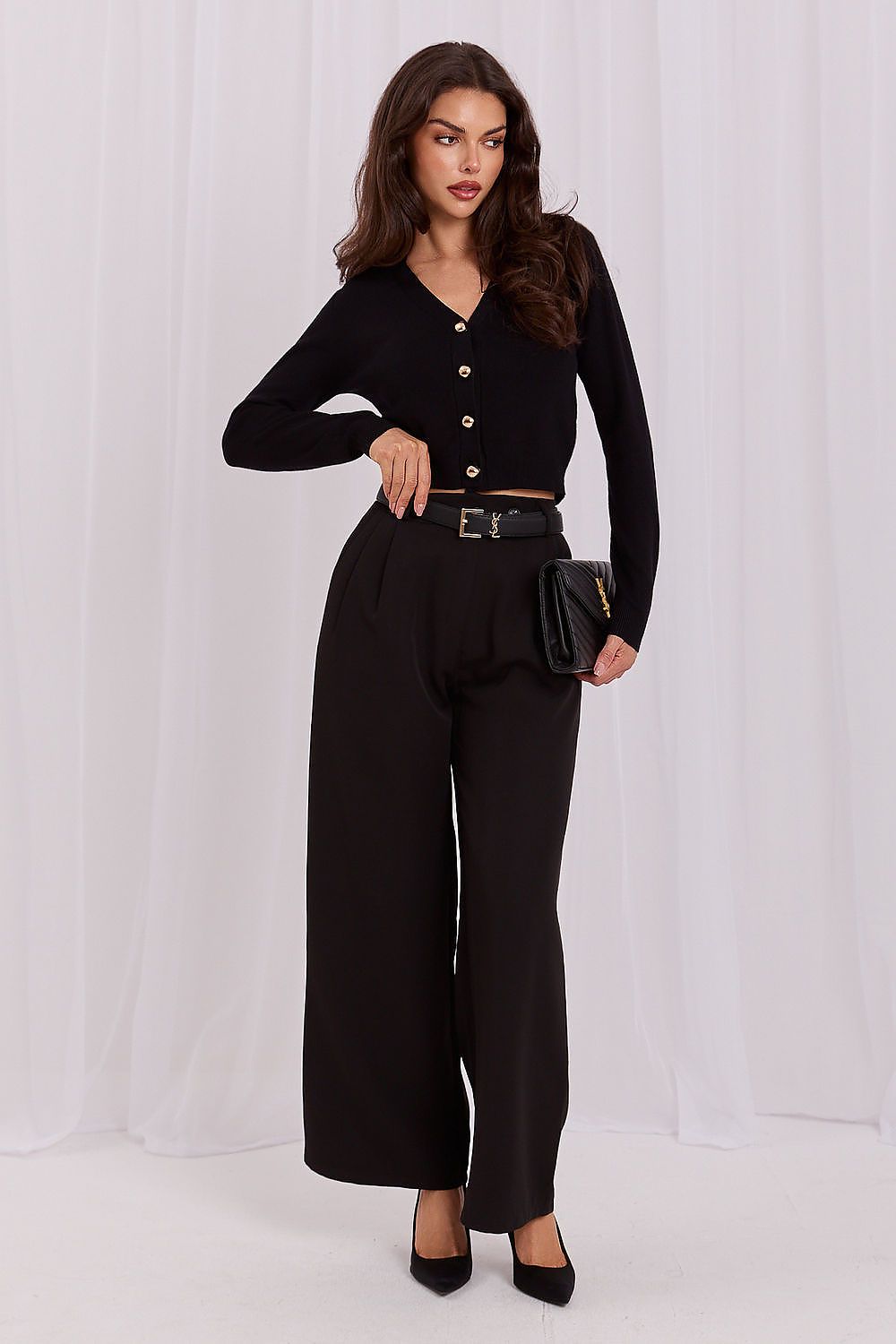 A woman modeling a black long-sleeve button-front top with gold buttons, paired with high-waisted black wide-leg trousers, a black belt, and black heels, standing against a white draped backdrop.