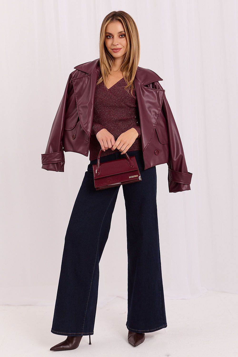 A woman models a dark burgundy faux-leather jacket over a matching burgundy top, paired with dark navy wide-leg jeans, brown pointed-toe heels, and a matching burgundy handbag against a white studio backdrop.