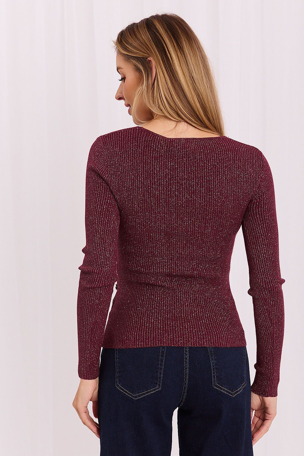 A woman shown from the back wearing a ribbed long-sleeve knit sweater in a deep burgundy shade, paired with dark blue jeans, against a light off-white curtain background.