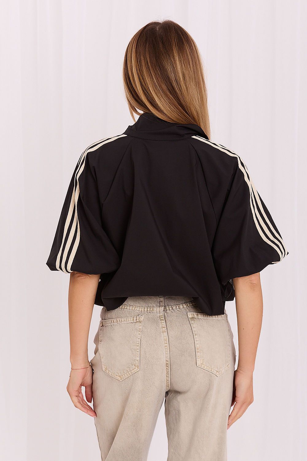 A woman shown from the back wearing a black cropped athletic jacket with three white stripes down the sleeves, paired with light beige jeans, against a white curtain background.
